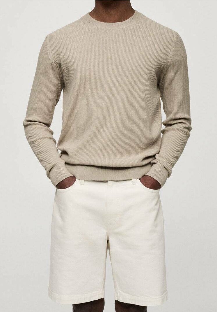 Homme portant un pull beige côtelé à manches longues et un short blanc cassé jusqu'aux genoux, mains dans les poches.