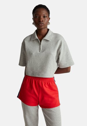 Šedý top na zip s krátkými rukávy a červené šortky. Šortky mají kontrastní modrý pruh podél stran. Hladká textura látky.