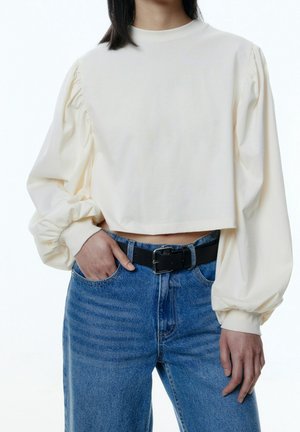 Vrouw die een cropped crème blouse draagt met geplooide lange mouwen en een high-waisted blauwe jeans met een zwarte riem, hand in de zak.