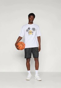 Weißes T-Shirt mit Grafik eines Palmenbaums und Basketball, kombiniert mit schwarzen Mesh-Shorts. Das Model hält einen orangefarbenen Basketball und trägt weiße Sneaker.
