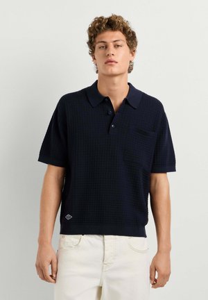 Junger Mann mit lockigem Haar, der ein dunkelblaues kurzärmeliges Strick-Poloshirt und off-white Hosen vor einem hellgrauen Hintergrund trägt.