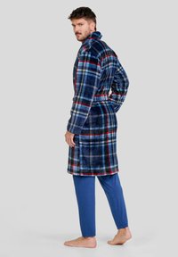 Bata de baño de felpa a cuadros en azul marino, rojo y azul claro, con cuello de chal y cinturón. Combinada con pantalones azules lisos, descalzo y en vista lateral.