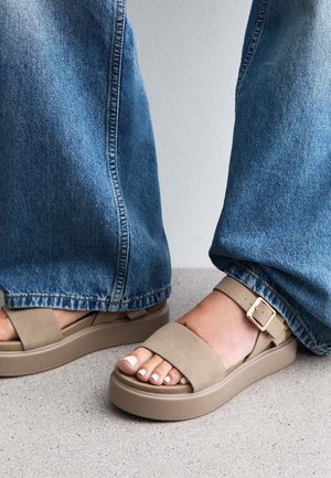 Sandales à plateforme beige avec de larges brides et un détail de boucle. Portées avec un jean en denim bleu ample, mettant en valeur le design et la couleur des chaussures.