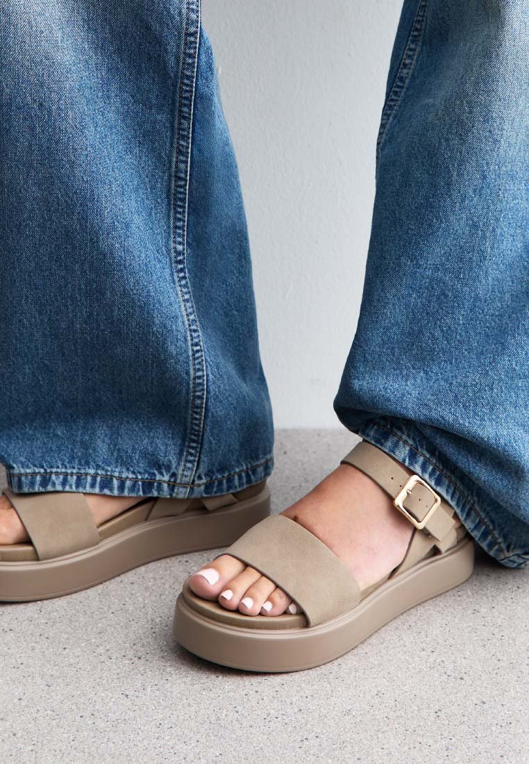 Sandali platform beige con ampia cinturino e dettaglio con fibbia. Indossati con jeans in denim blu larghi, evidenziando il design e il colore delle scarpe.