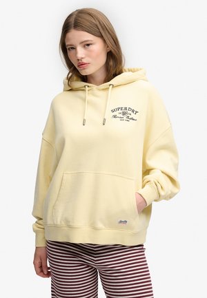 Ung kvinde iført en lysegul Superdry-hættetrøje med frontlomme og bordeaux og hvide stribede bukser, stående mod en hvid baggrund.