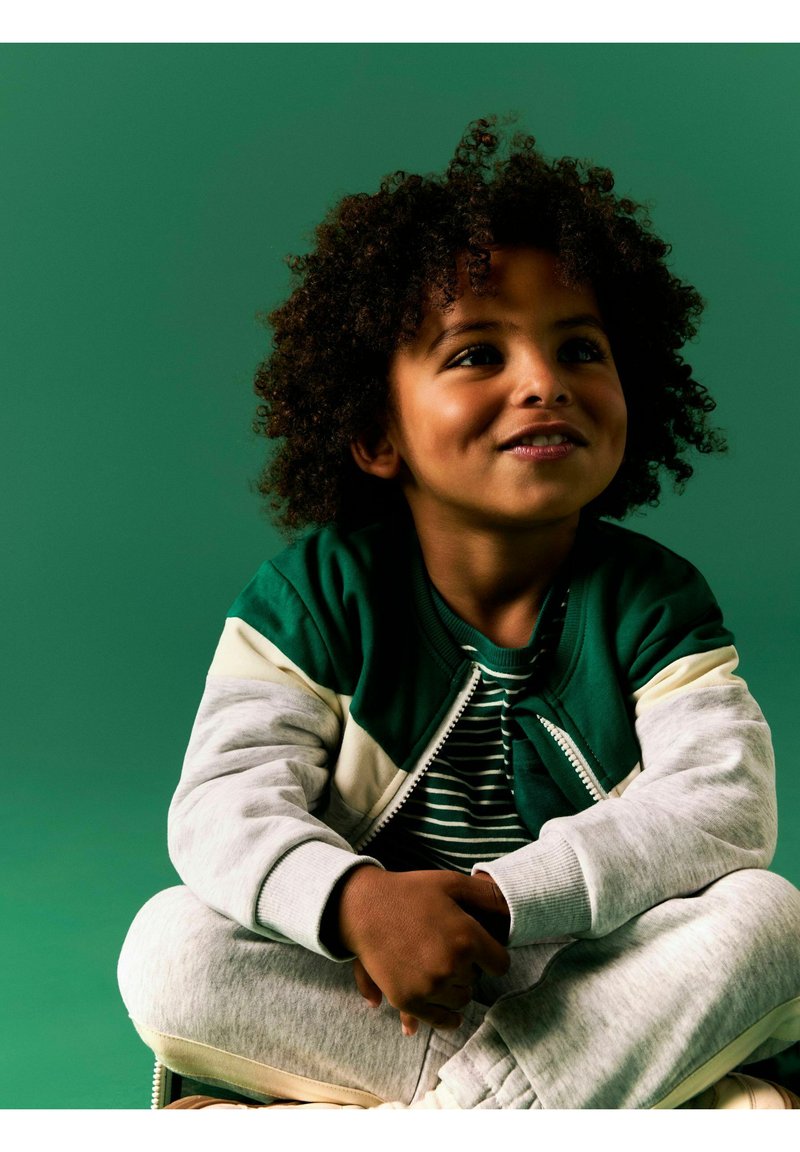 Jeune enfant aux cheveux bouclés assis en tailleur et souriant, portant une veste verte et blanche sur une chemise rayée, sur fond vert.
