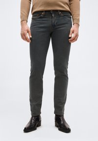 Άνδρας με στενά, σκούρα γκρι τζιν, ανοιχτό καφέ πουλόβερ και μαύρα μποτάκια αστραγάλου, που στέκεται σε απλό, ανοιχτό φόντο.