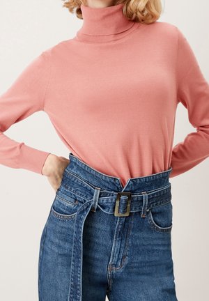 Donna che indossa un maglione dolcevita rosa infilato in jeans di denim blu a vita alta con una cintura larga e fibbia rettangolare.