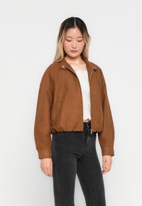 Veste courte marron avec une texture lisse, fermeture éclair et un col simple, associée à un jean noir. Design classique avec un minimum de détails.