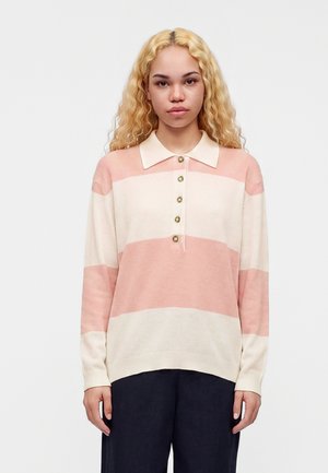 Jeune femme aux longs cheveux blonds bouclés portant un polo à manches longues rayé rose pâle et crème et un pantalon foncé, debout devant un fond blanc.