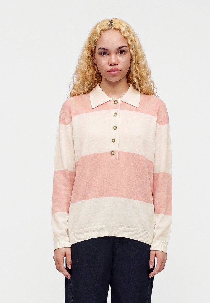 Jeune femme aux longs cheveux blonds bouclés portant un polo à manches longues rayé rose pâle et crème et un pantalon foncé, debout devant un fond blanc.