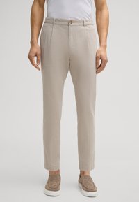 Pantalon beige clair en tissu texturé, avec fermeture par bouton, poches latérales et jambes fuselées, associé à des chaussures à enfiler.