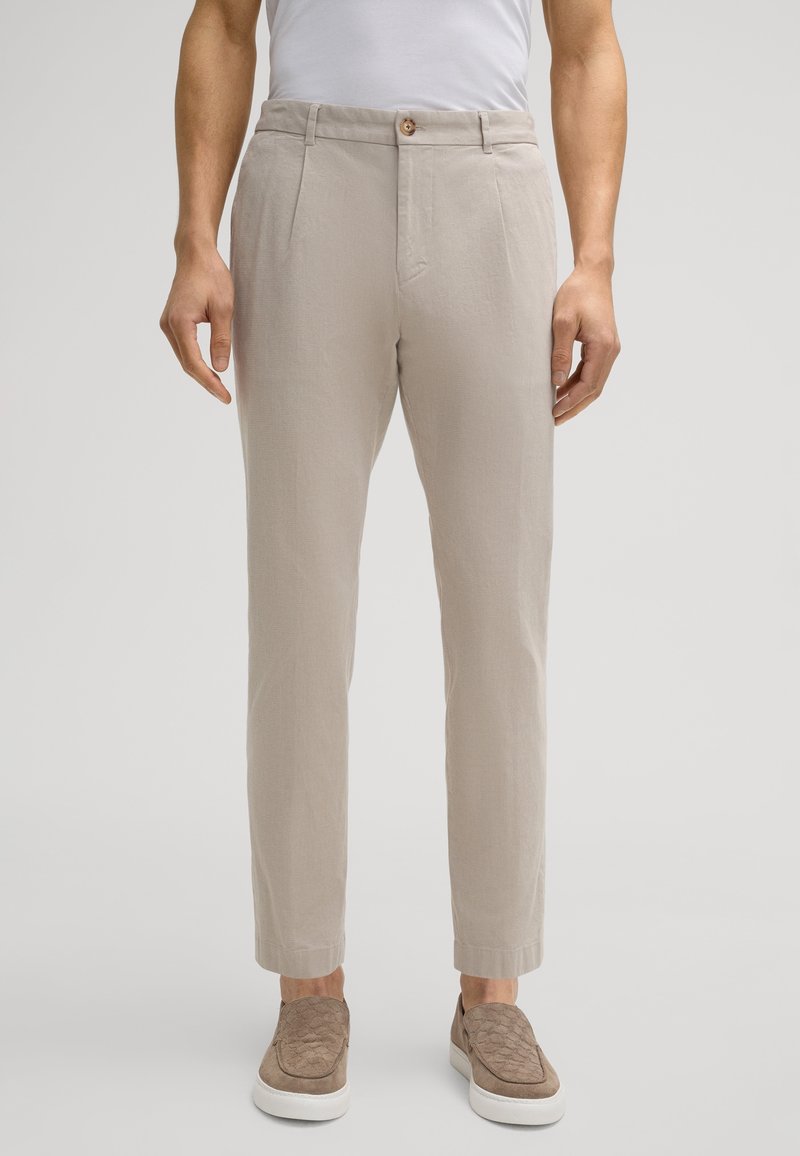 Pantalon beige clair en tissu texturé, avec fermeture par bouton, poches latérales et jambes fuselées, associé à des chaussures à enfiler.