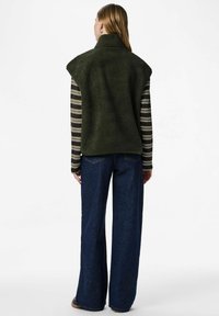 Sieviete ar vidēja garuma matiem valkā tumši zaļu fliša veste pār svītrainu garām piedurknēm kreklu un platām zilo džinsu biksēm, stāvoša ar muguru pret skatītāju.