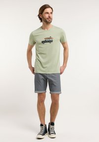 T-shirt verde chiaro con una grafica di un'auto che trasporta un kayak, abbinato a pantaloni corti grigi arrotolati e sneakers alte nere.