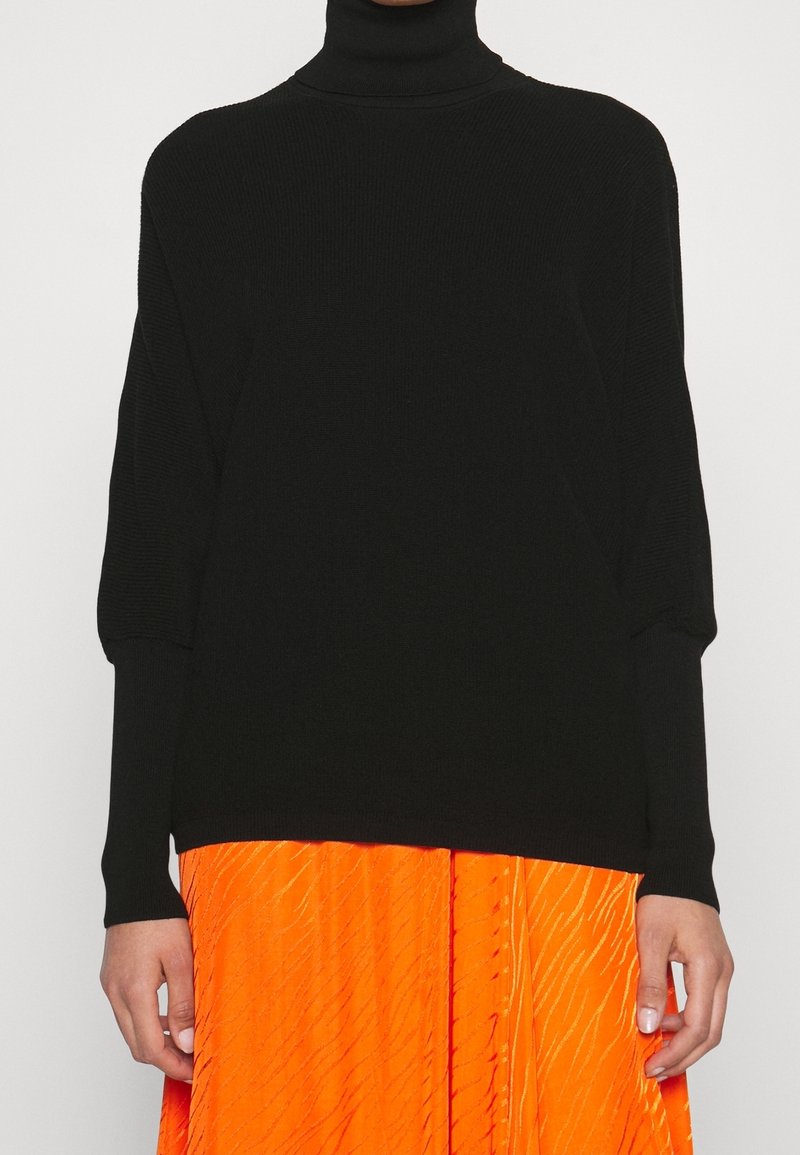 Pull à col roulé noir côtelé à manches longues, associé à une jupe plissée orange. Le tissu semble doux et légèrement texturé.