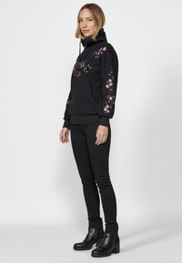 Sweat-shirt à capuche noir avec broderies florales sur les manches, surface texturée, associé à un jean skinny noir et des bottines à talons épais.