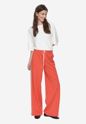 Czerwone spodnie o szerokich nogawkach z bocznymi paskami i troczkiem w talii, zestawione z białym t-shirtem o luźnym kroju. Gładka faktura tkaniny.