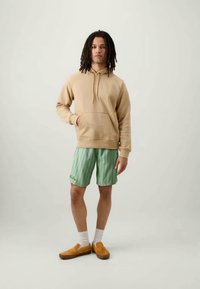 Beigā kapucē ar priekšējo kabatu, apvienota ar zaļām svītrainām šortiem. Brūni slip-on apavi un baltas zeķes noslēdz šo apģērbu.