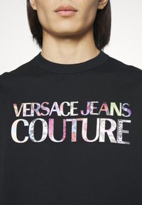 Detailní záběr osoby v černém tričku s nápisem "VERSACE JEANS COUTURE" v barevném květinovém vzoru přes hruď.