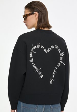 Frau mit kurzen braunen Haaren, die Schildpatt-Sonnenbrillen trägt und einen schwarzen Pullover mit weißem Schriftzug in Herzform auf dem Rücken.