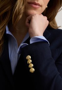Nærbillede af en person iført en navy blazer med guldknapper og en lyseblå skjorte, der hviler hagen på sin hånd.