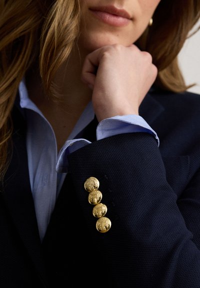 Gros plan d'une personne portant un blazer bleu marine avec des boutons dorés et une chemise bleu clair, posant son menton sur sa main.