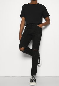 Camiseta negra, vaqueros negros rasgados con adornos, ajustados, y zapatillas altas negras. Diseño sencillo con acentos de tela ausentes.