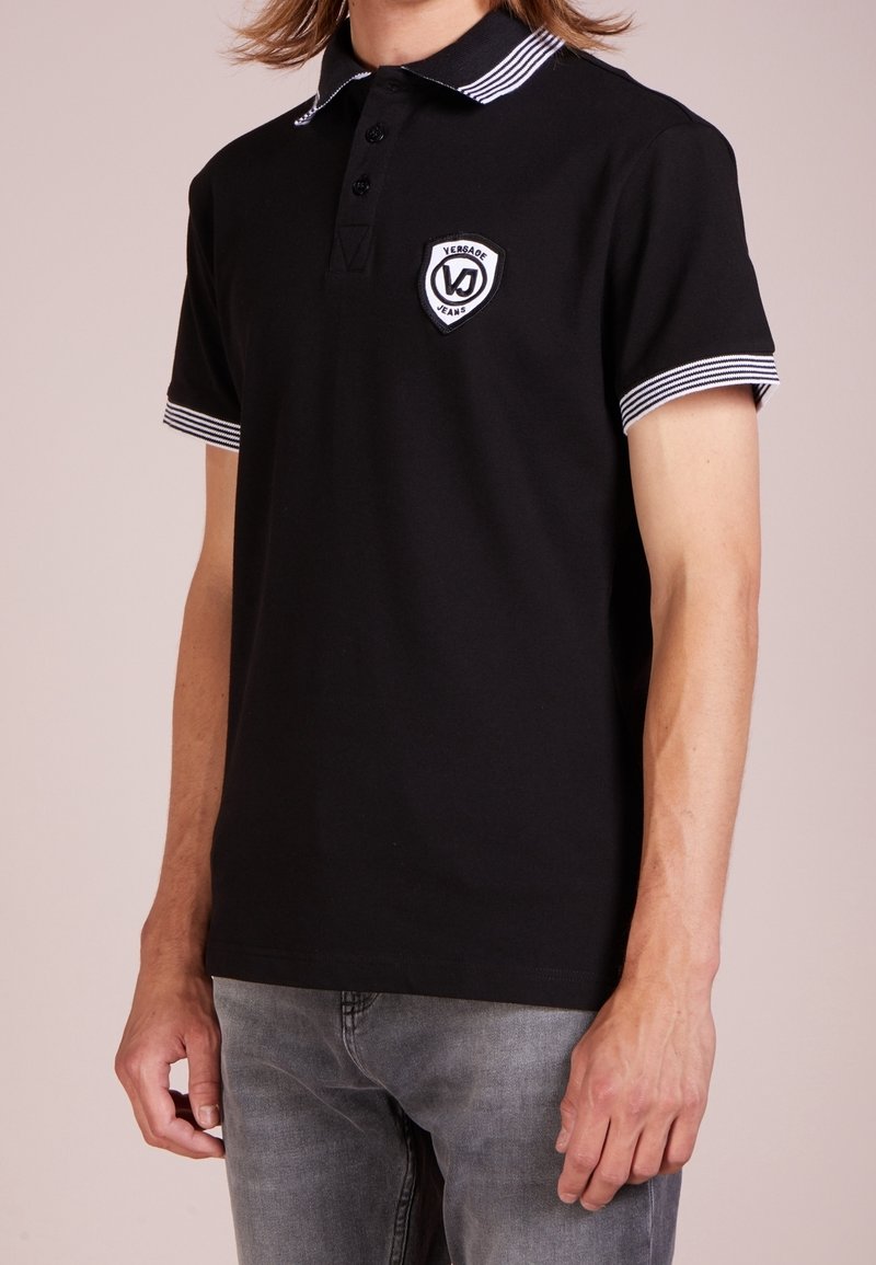 Homme portant un polo noir avec col et manches rayés blancs, logo Versace Jeans sur la poitrine, associé à un jean gris.