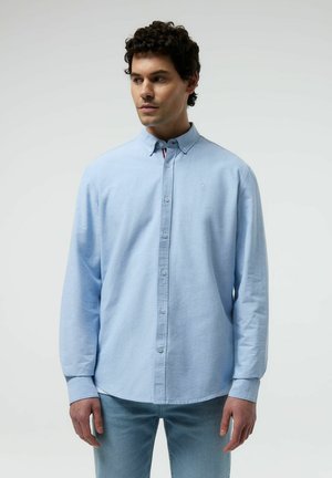 Mężczyzna o kręconych ciemnych włosach, ubrany w jasnoniebieską koszulę na guziki i niebieskie dżinsy, patrzący lekko w lewo na tle zwykłego tła.