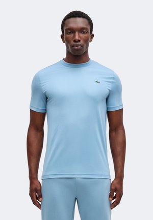 Homme portant une chemise bleu clair à manches courtes et un pantalon assorti, debout face à l'avant sur un fond uni.