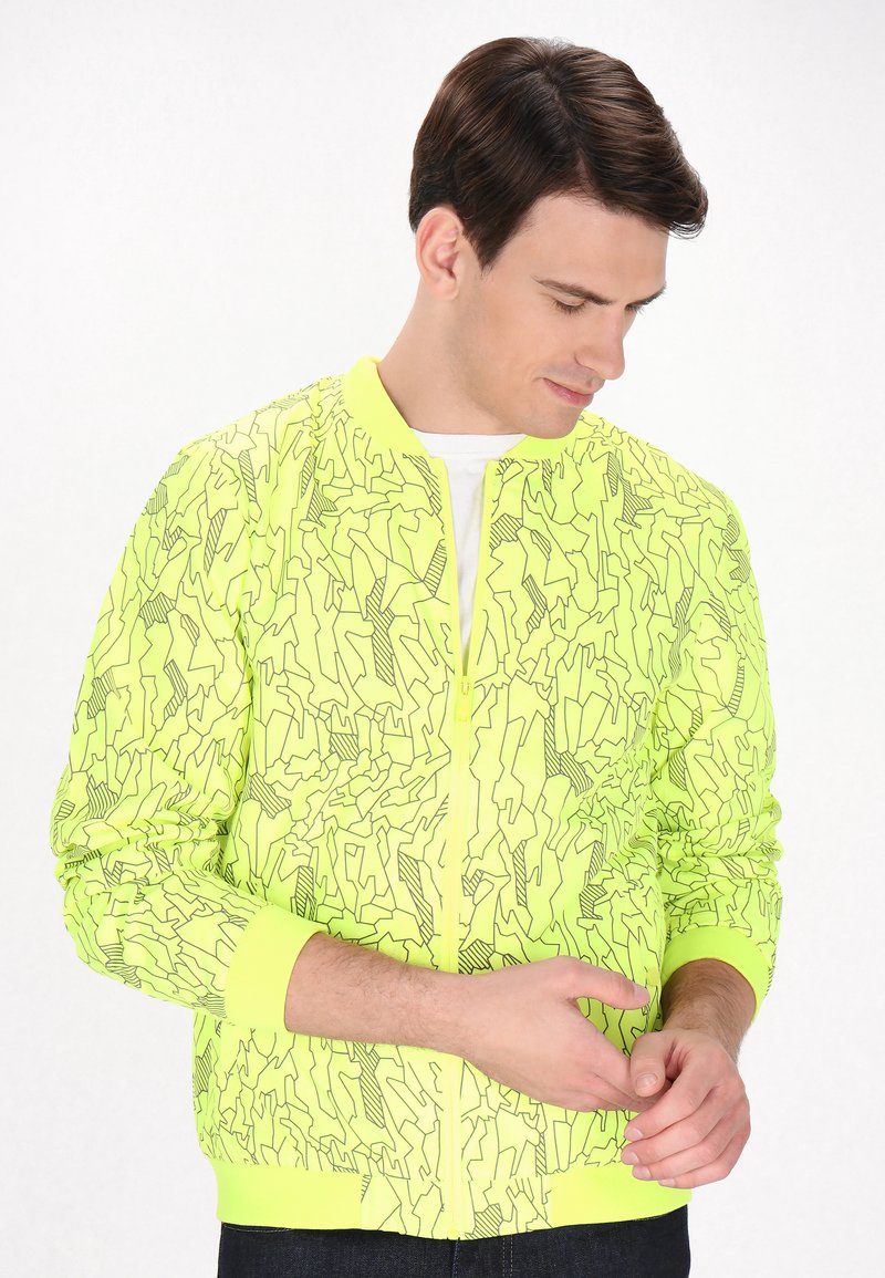 Jachetă bomber galben neon cu modele abstracte din linii negre, mansete și tiv cu nervuri. Material neted, închidere cu fermoar pe față, croială lejeră.