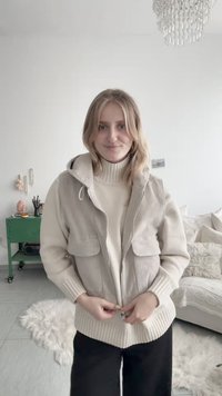 Ung kvinde med blondt hår iført en beige jakke over en cremefarvet rullekrave, stående i en moderne stue med en grøn reol og en hvid sofa.