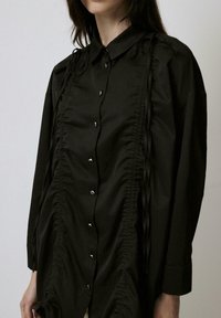 Black button-up shirt made of smooth fabric features ruching down the front, with thin drawstrings and metallic buttons along the closure.