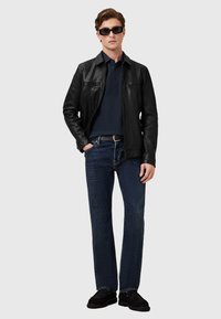 Schwarze Lederjacke, navy Polohemd, dunkle Jeans, schwarze Schuhe. Sonnenbrille und ein Gürtel runden den Look ab. Lässig, moderne Passform.