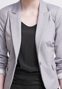 Jasnoszary żakiet o zbudowanej formie, z klapami w kształcie litery notch i subtelną teksturą. Posiada dwie przednie kieszenie i zapięcie na guziki. Noszony na czarnym topie.