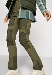 Khaki-Hosen zum Wandern mit zweifarbiger Panelgestaltung und Reißverschlusstaschen, kombiniert mit hellen Sportschuhen, vor einem neutralen Hintergrund.