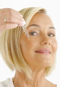 Transparent dråpeflaske med rosa hette holdt over en persons ansikt, som slipper ut en klar væske på huden. Kort, blondt hår og nøytral bakgrunn.