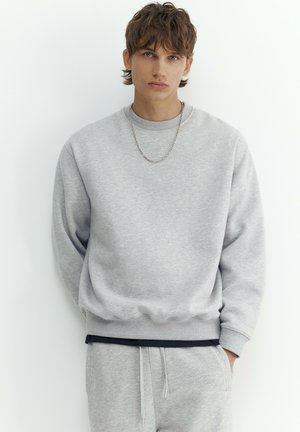 Jeune homme aux cheveux bruns courts portant un sweat-shirt gris clair et un pantalon de survêtement assorti, debout sur un fond blanc.