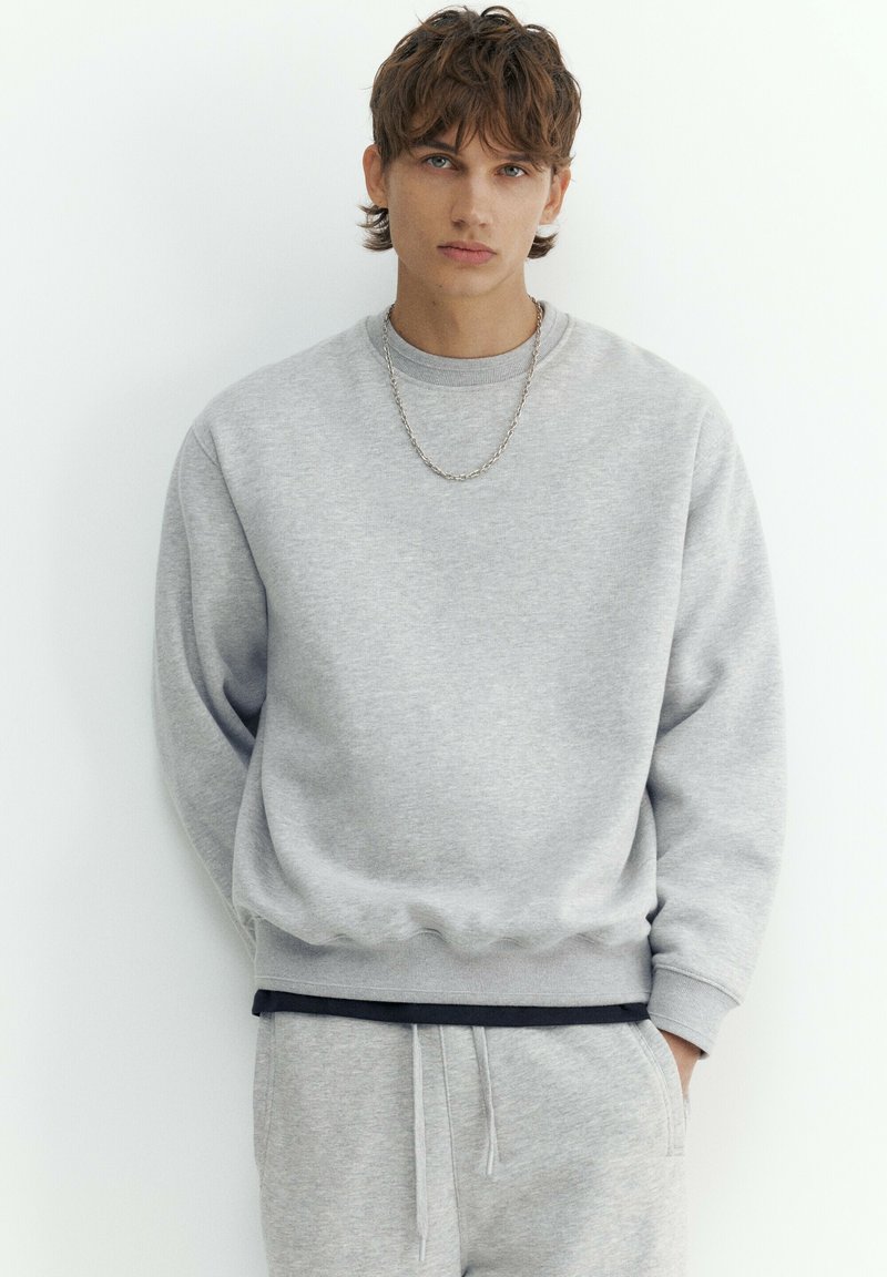 Jeune homme aux cheveux bruns courts portant un sweat-shirt gris clair et un pantalon de survêtement assorti, debout sur un fond blanc.