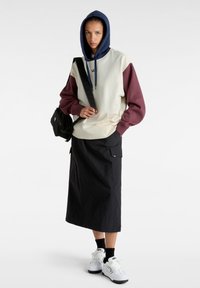Felpa con cappuccio di colore crema con maniche bordeaux, abbinata a una lunga gonna nera e sneakers bianche, con una borsa a spalla.