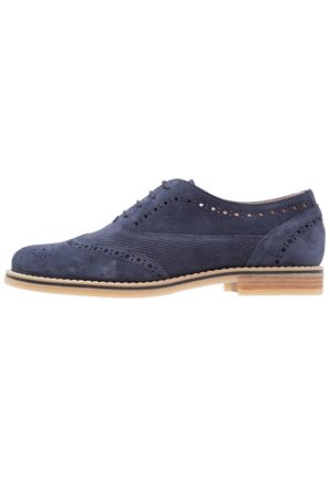 Marineblå ruskindssko med brogue-detaljer, perforeret mønster, tekstureret sidelæder, beige sål, kort stablet træhæl og snørelukning.