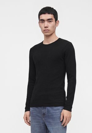 Joven con el pelo corto y negro que lleva una camiseta negra lisa de manga larga y vaqueros azules, de pie sobre un fondo blanco.