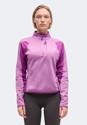 Femme portant un haut violet à manches longues avec une fermeture éclair partielle et un motif texturé, ainsi qu’un legging noir, debout devant un fond uni.