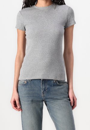 Femme portant un t-shirt gris côtelé ajusté à manches courtes et un jean bleu, debout devant un fond blanc.