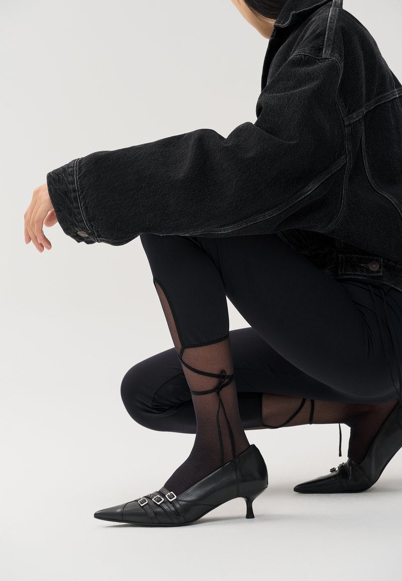 Zwarte kitten-heel schoenen met spitse neuzen en drie metalen gespen. Het model draagt zwarte doorzichtige panty's en zit in een spijkerjack.