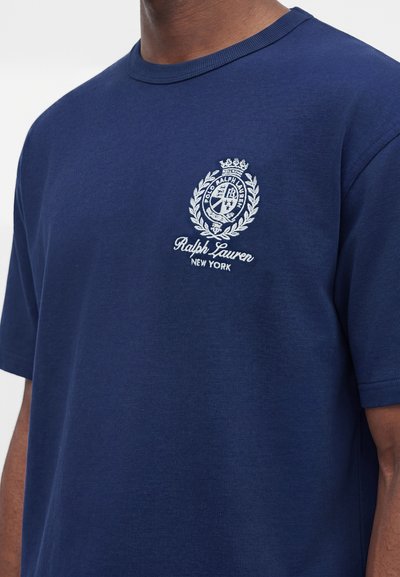 Άνδρας που φοράει ναυτικό μπλε μπλουζάκι Ralph Lauren με λευκό κεντημένο έμβλημα και το κείμενο «Ralph Lauren New York» στο στήθος.