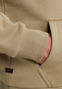 Beige Sweatshirt mit geripptem Bündchen und Seitentasche. Verfügt über ein Markenetikett an der unteren linken Seite und eine glatte Stoffstruktur.