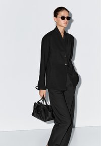 Vrouw die een zwarte zonnebril, een losse zwarte blazer en broek draagt, en een zwarte leren handtas vasthoudt tegen een lichte achtergrond.