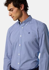 Camisa de cuadros en azul y blanco con cuello clásico, que presenta botones blancos y un pequeño logo en el pecho.